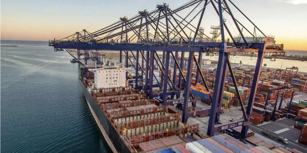 Aerial view of a container terminal at sunset, with ship-to-shore gantry cranes over a loaded container vessel.